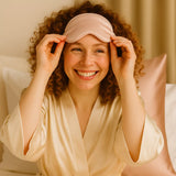 Femme souriante aux cheveux bouclés mettant un masque de nuit en soie rose, ambiance douce du soir, taie d’oreiller assortie en arrière-plan.